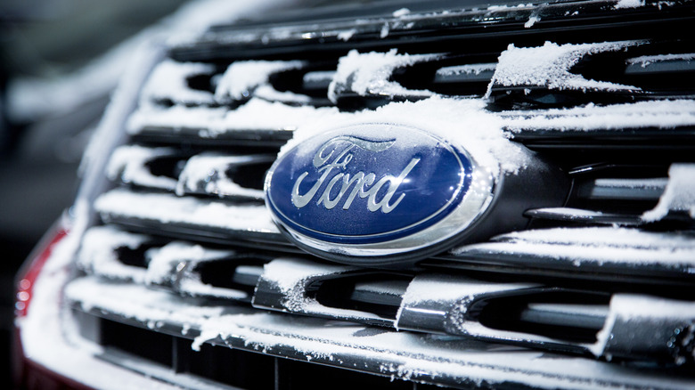 Um carro de teste camuflado em uma pista de gelo, com edifícios e neve ao fundo.