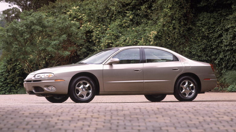 Oldsmobile Aurora 1ª geração estacionado em um gramado com plantações ao fundo