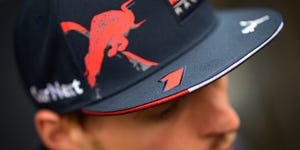 Max Verstappen sorrindo, vestindo uniforme de corrida e capacete, com um carro de corrida ao fundo
