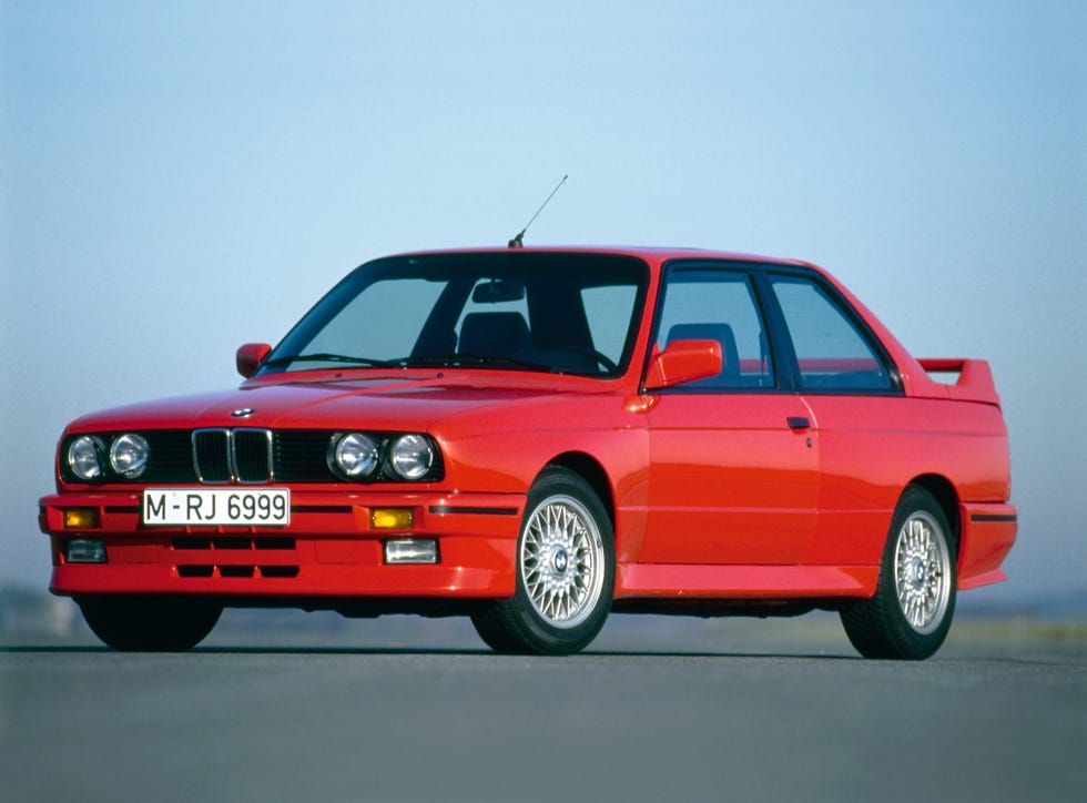BMW M3 E30 cinza estacionado em ambiente urbano com prédio de tijolos ao fundo